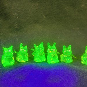 Puede incluir: Una colección de seis pequeñas figuras verdes translúcidas, cada una con una forma estilizada de animal. Las figuras están espaciadas uniformemente y parecen estar hechas de un material brillante. El fondo es una tela oscura y texturizada.