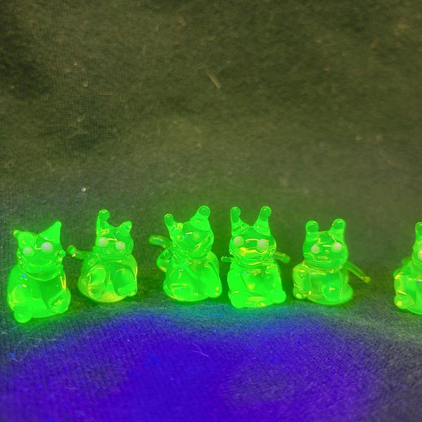 May include: A collection of six small, translucent green figurines, each with a stylized animal-like form. The figures are evenly spaced and appear to be made of a glowing material. The background is a dark, textured fabric.