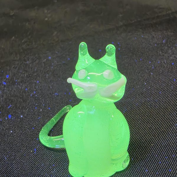 May include: A small, translucent, neon green glass cat figurine. The cat features two pointed ears, two small white circular eyes, and a prominent white mustache across its face. A curved glass tail extends from the side of the rounded body.
