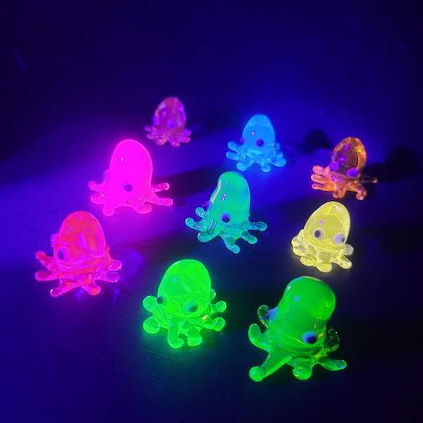 May include: A collection of nine small, translucent octopus figurines made of glass. Each figurine glows in vibrant neon colors including pink, green, yellow, and orange under ultraviolet light. The octopuses feature rounded heads and multiple curled tentacles, with distinct dark eyes visible on each creature.