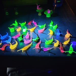 Puede incluir: Una colección de figuras brillantes con forma de babosa en varios colores, incluyendo verde, amarillo, rosa y azul. Las figuras están dispuestas sobre una superficie oscura, iluminadas por luz negra.