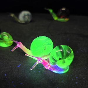 Puede incluir: Varias figuras de caracoles de vidrio con acentos verdes, rosas y azules brillantes se disponen sobre un fondo oscuro. Los caracoles tienen cuerpos y antenas detallados. La imagen está bien iluminada, resaltando los colores vibrantes y los intrincados detalles del arte en vidrio.