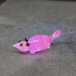 Puede incluir: Una pequeña figura de ratón de cristal hecha a mano. El ratón es principalmente rosa con vetas blancas, y presenta ojos oscuros y pequeñas orejas negras. La cola es transparente. La figura mide aproximadamente 5 cm de largo.