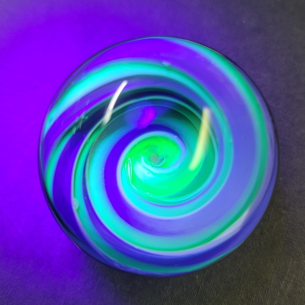May include: A glass marble with a swirling pattern of green, white, and blue. The marble is illuminated with a UV light, causing the green to glow.
