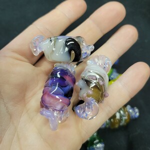 May include: A collection of colourful glass candy-shaped ornaments. The ornaments are made of clear glass with swirls of purple, pink, white, black, and brown.