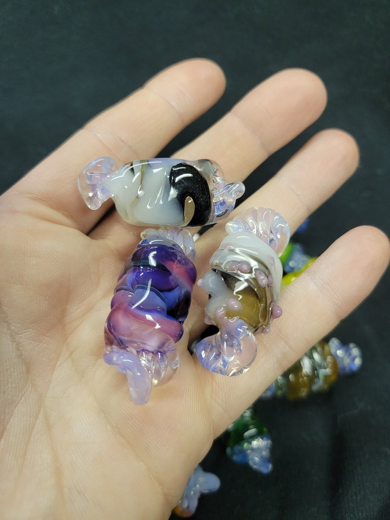 May include: A collection of colourful glass candy-shaped ornaments. The ornaments are made of clear glass with swirls of purple, pink, white, black, and brown.
