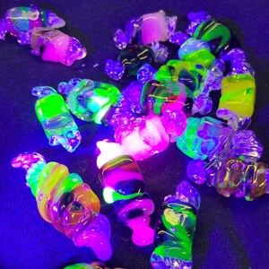 May include: A collection of colourful glass beads, some with swirls and others with a smooth finish, glow brightly under a black light. The beads are a mix of green, pink, yellow, and black.