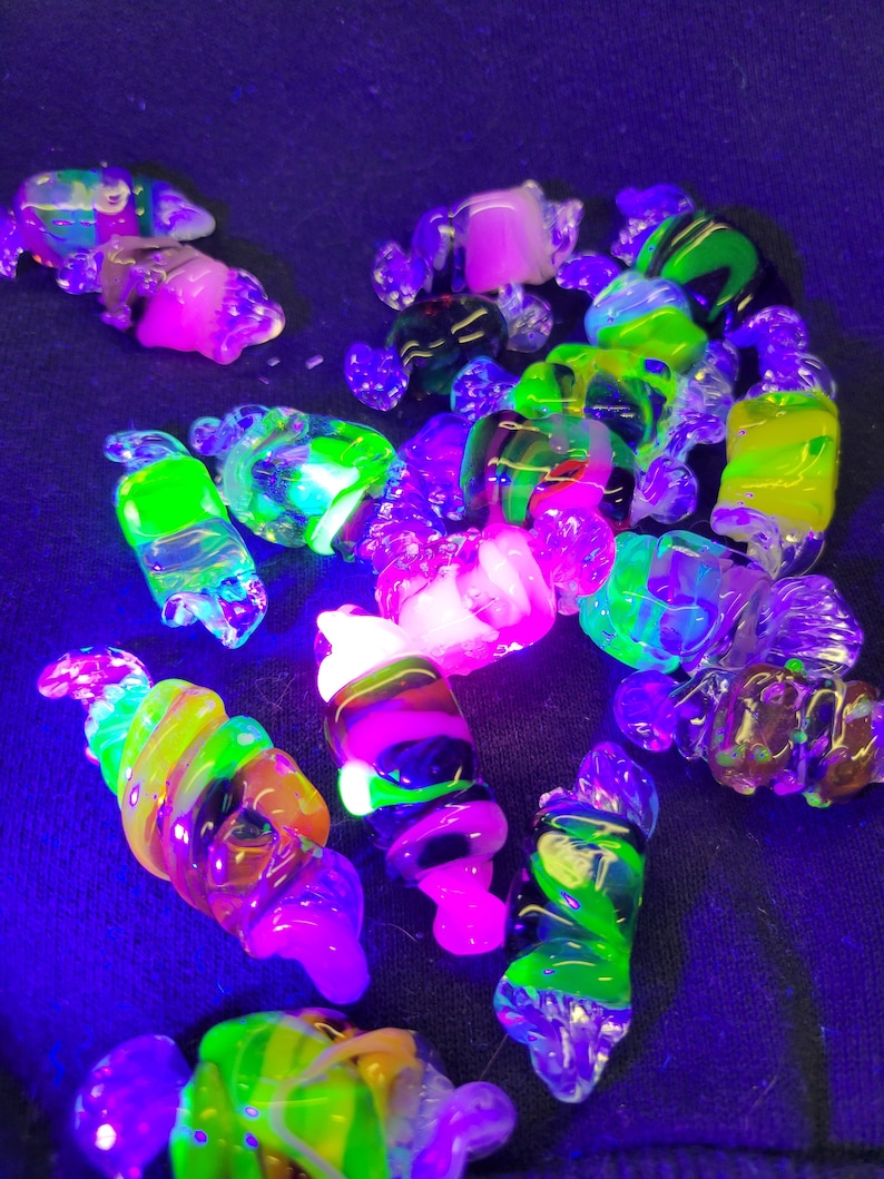 May include: A collection of colourful glass beads, some with swirls and others with a smooth finish, glow brightly under a black light. The beads are a mix of green, pink, yellow, and black.