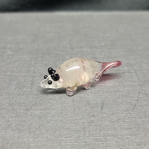 Puede incluir: Una pequeña figurita de vidrio de un ratón rosa y blanco con ojos negros. El ratón está sentado sobre una superficie gris.