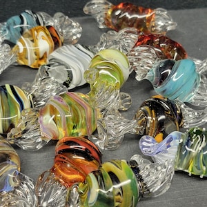 Puede incluir: Una colección de esculturas de caramelo de vidrio de colores. Cada pieza está soplada a mano y presenta un patrón de remolino único. Las esculturas de caramelo están hechas de vidrio transparente con remolinos coloridos de azul, verde, rojo, amarillo y naranja.