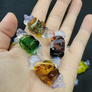 May include: Five glass candy charms in various colours and patterns, including green, brown, white, and blue. The charms are shaped like wrapped sweets.