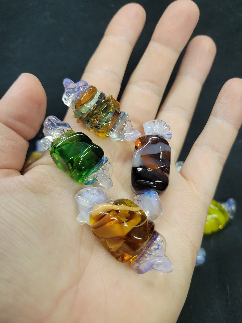 May include: Five glass candy charms in various colours and patterns, including green, brown, white, and blue. The charms are shaped like wrapped sweets.