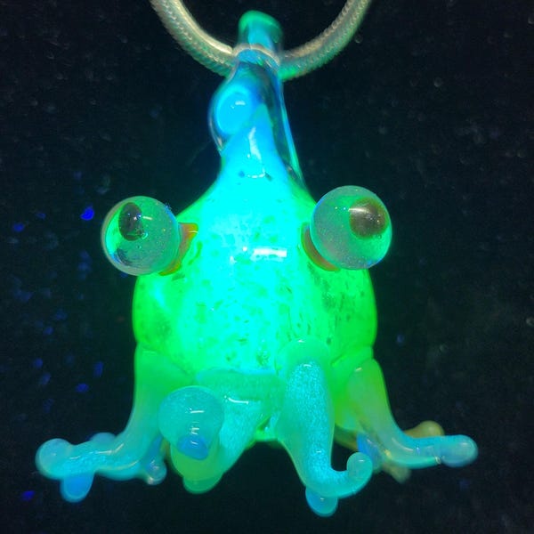 May include: A glowing glass pendant in the shape of a whimsical creature, with large, round eyes and a light green body. The pendant is suspended from a silver chain, and the creature's body glows with a bright, neon green color.