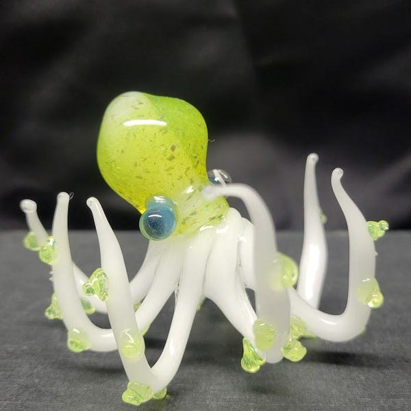 May include: A glass octopus sculpture with a yellow and white color scheme. The head is yellow with green speckles, and the eyes are blue. The tentacles are white with green accents. The sculpture is on a dark gray surface.