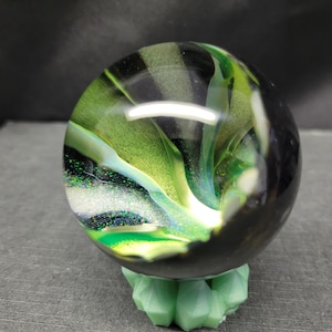May include: A clear glass marble with swirling green and black colors, resting on a light green geometric stand. The marble has a reflective surface, showcasing the internal design. The stand is a light green color.