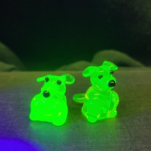 May include: Two small, bright green glass figurines of dogs. The dogs are sitting with their heads turned to the side. The figurines are made of uranium glass, which glows under a black light.