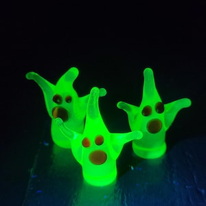 以下が含まれることがあります： オレンジ色の目と口を持つ、3つの光る緑色のゴーストフィギュア。フィギュアは腕を広げており、暗い背景に配置されています。オブジェクトは半透明の素材でできているようです。