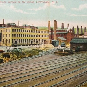 May include: A vintage postcard depicting a large industrial complex with multiple buildings and smokestacks. The text on the postcard reads "Corning Glass Works, largest in the world, CORNING, N.Y."