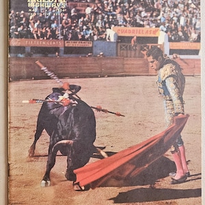 Pode incluir: Capa de revista vintage com uma cena de tourada. A capa diz "Arizona Highways" e a data "Maio de 1954". Um toureiro com uma capa vermelha enfrenta um touro preto numa arena de areia, com uma multidão ao fundo.