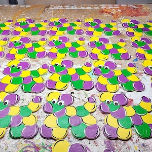 May include: A collection of colorful, wooden Mardi Gras-themed decorations. Each piece is a stylized dog shape, painted in purple, green, and yellow. The decorations are arranged on a wooden surface.