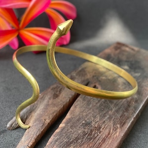 Puede incluir: Un brazalete de serpiente de oro con un extremo puntiagudo. El brazalete está apoyado en una pieza de madera oscura.