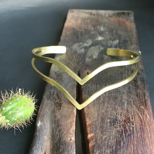 May include: A gold-toned double-layered metal cuff bracelet with a V-shaped design. The bracelet is displayed on a piece of weathered wood.