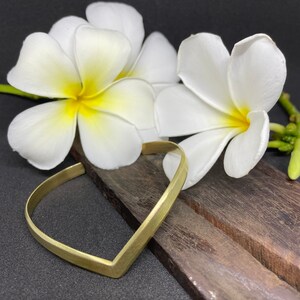 May include: A gold-toned, V-shaped bangle bracelet resting on a wooden surface with white plumeria flowers.