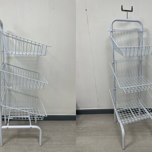 May include: White metal three-tiered display stand with wire baskets. The stand has a hook at the top and a base with three legs.