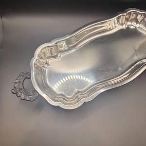 Könnte beinhalten: Ein längliches silberfarbenes Serviertablett mit gewelltem Rand und reflektierender Metalloberfläche. Das Tablett verfügt über einen einzelnen dunklen, verzierten, schnörkelartigen Griff an einem Ende, der einen Kontrast zum polierten Silberkörper bildet.