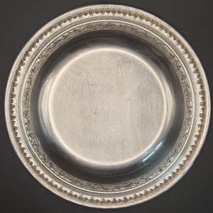 May include: A round silver-toned metal bowl featuring a decorative embossed border with a repeating scroll and bead pattern. The center of the bowl displays faint engraved text that reads Reed & Barton 1201, with a separate marking of 1475 below it.
