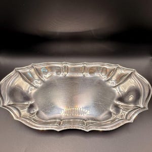 May include: An oval silver-toned serving tray with a polished, reflective surface and a scalloped, multi-faceted rim. The tray features a smooth, slightly concave center and a decorative, undulating edge that creates a distinct border.