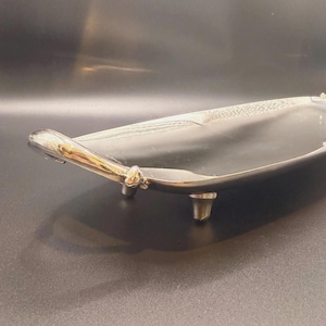 Puede incluir: Una bandeja de metal plateado de forma ovalada y alargada con una superficie interior lisa y reflectante. La bandeja cuenta con dos asas curvas en los extremos y se apoya sobre cuatro patas pequeñas y resistentes, lo que le da un aspecto elevado.