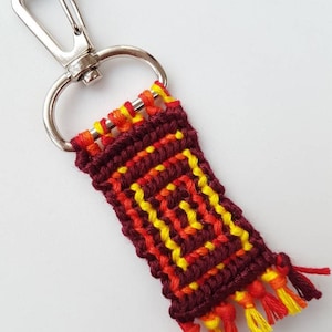 May include: A maroon, yellow, and orange woven keychain with a silver metal clasp. The keychain has a fringe on the bottom.