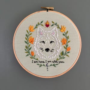 Puede incluir: Un bastidor de bordado circular con tela clara que presenta un rostro de lobo blanco bordado a mano, rodeado de flores naranjas y hojas verdes. Sobre el lobo hay un pequeño corazón rojo con llamas. El texto dice: I am here. I am with you.