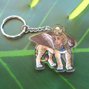 May include: A gold and brown acrylic keychain featuring a winged lion with a human head wearing a crown and jewellery. The keychain has a silver metal ring.
