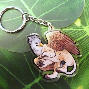 May include: A clear acrylic keychain featuring a detailed illustration of a griffin, a mythical creature with the head and wings of an eagle and the body of a lion. The griffin is depicted in a crouching pose with its wings spread out.