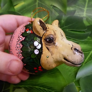 Op de afbeelding: Een hand houdt een roze en groene kerstbal vast in de vorm van een kamelenkop. De bal is versierd met hulst en bessen.