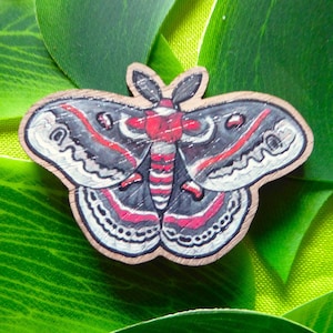 May include: A wooden moth-shaped pin with a detailed design. The moth features gray wings with red and white accents, a red body, and a light brown border. The pin is set against a backdrop of green leaves.