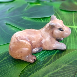 Puede incluir: Figurita de capibara de cerámica en tonos marrones y beige, con detalles negros en las patas y los ojos. La figura está tumbada sobre un lecho de hojas verdes grandes, creando un entorno naturalista. Escultura en miniatura detallada.