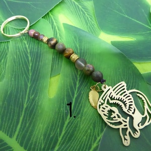May include: Gold key chain with a winged cat charm and a beaded strand with tiger's eye, pink, and green stones. The number "1." is visible in the background.