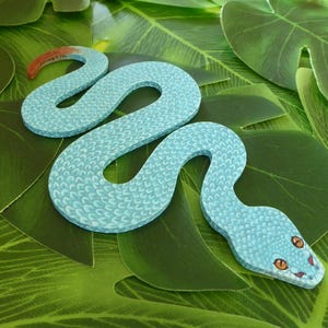 May include: A light blue snake with a textured pattern, curled up on a green leafy background.
