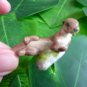 May include: A miniature ceramic otter figurine, lying on its back with its paws in the air. The otter is brown and tan with a glossy finish. It rests on a small, green and white base, set against a backdrop of green leaves.