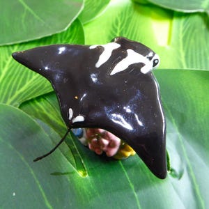 May include: A glossy black ceramic manta ray figurine with white markings, set against a backdrop of large green leaves. The figurine has a detailed design and is a decorative item.