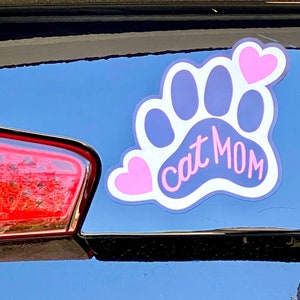 May include: A white paw print with a pink heart on each side and the text "Cat Mom" in pink lettering.