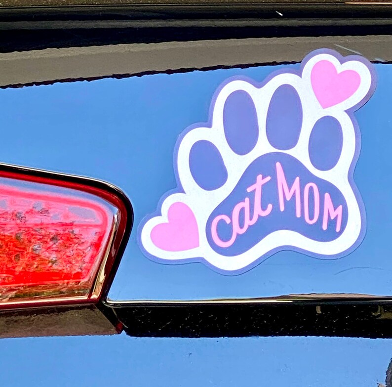 May include: A white paw print with a pink heart on each side and the text "Cat Mom" in pink lettering.