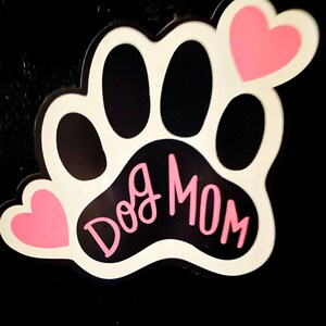 May include: White paw print with black paw pads and the text "Dog Mom" in pink lettering. Two pink hearts are on either side of the paw print.