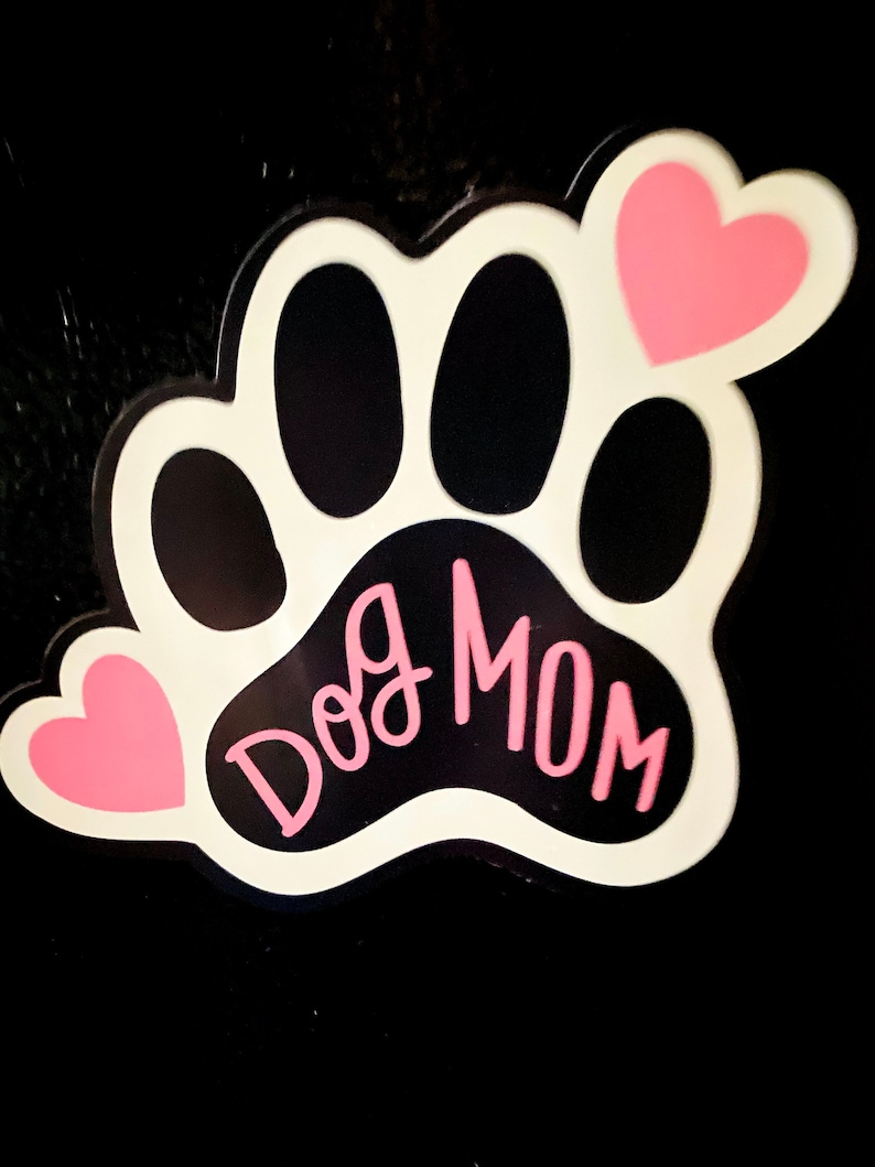 May include: White paw print with black paw pads and the text "Dog Mom" in pink lettering. Two pink hearts are on either side of the paw print.