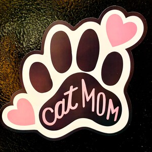 May include: A white paw print with black paw pads and the text "cat mom" in pink lettering. Two pink hearts are on the paw print.