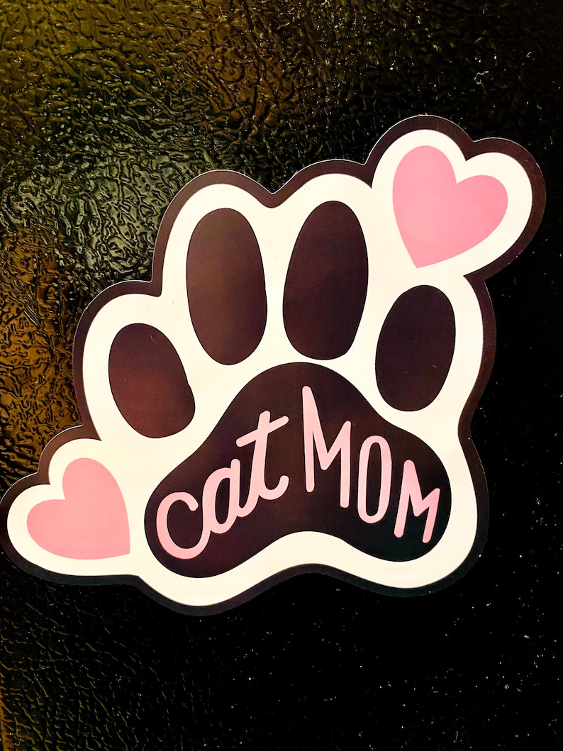 May include: A white paw print with black paw pads and the text "cat mom" in pink lettering. Two pink hearts are on the paw print.