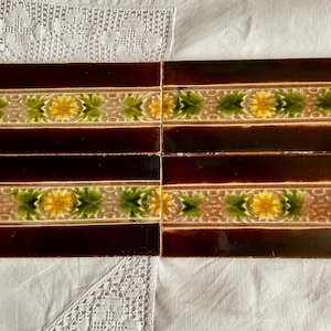 May include: Six rectangular ceramic tiles with a dark brown background and a decorative border. The border features a repeating pattern of yellow flowers and green leaves on a beige background. The tiles are arranged in two rows of three.